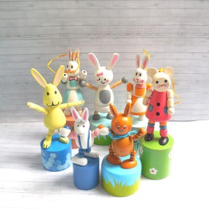 May include: A collection of wooden bunny and mouse figurines with articulated limbs. The bunnies are painted in various colors, including yellow, white, and orange, and stand on colorful cylindrical bases. The mouse wears a pink dress. Each figurine has a unique outfit.