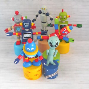 May include: A collection of six wooden push-up toys featuring colorful robots and a green alien. Each figure is mounted on a cylindrical base with various patterns, including planets and stars. The robots display diverse designs with red, blue, green, and grey paint, featuring articulated limbs and painted facial expressions.