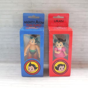 May include: Two vintage boxed push puppets. One blue box features "Mighty Atom" with a figure in a swimsuit. The other red box features "Uran" with a figure in a pink dress. Both boxes have a circular logo with a character's face.