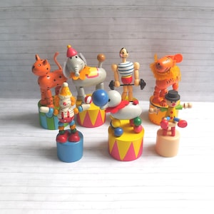 May include: A collection of colorful wooden circus-themed toys, including a tiger, elephant, clown, and strongman. Each figure is mounted on a small, painted platform. The toys are painted in bright colors and feature whimsical details.