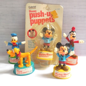 May include: A collection of vintage Gabriel Disney push-up puppets featuring Mickey Mouse, Donald Duck, Pluto, and Goofy. The figures are mounted on colourful circular bases with labels. One Mickey Mouse puppet remains in its original cardboard packaging.