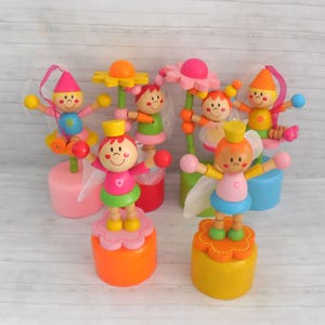 May include: A collection of six wooden spring-loaded toys featuring colorful figures with painted faces, conical hats, and flower-shaped bases. Each figure has outstretched arms and is mounted on a cylindrical stand in bright shades of pink, orange, and yellow.