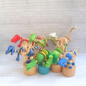 May include: A collection of five articulated wooden dinosaur figures in red, green, blue, and natural wood tones. Each dinosaur is mounted on a cylindrical wooden base, featuring segmented bodies, movable limbs, and distinct head shapes.