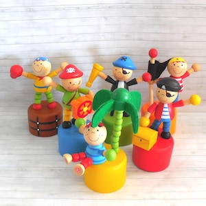 May include: A collection of colorful wooden pirate-themed toys. Each toy features a smiling face, a unique pirate outfit, and is mounted on a brightly colored base. Accessories include a treasure chest, telescope, and palm tree.