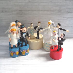 May include: A set of wooden wedding-themed puppet figures. The puppets are dressed in traditional wedding attire, including a white dress, a black tuxedo, and a veil. The puppets are standing on wooden bases with blue and red paint.