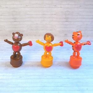May include: Three bobblehead toys with red hearts on their chests. The toys are a brown monkey, a yellow lion, and an orange tiger. The toys are all standing on a wooden surface.