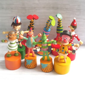 Op de afbeelding: Een verzameling kleurrijk houten speelgoed, waaronder een aap, een clown en andere grillige figuren. Elke figuur staat op een cilindrische basis en heeft beweegbare armen en een uniek ontwerp. Het speelgoed is geschilderd in heldere kleuren en heeft een vintage esthetiek.