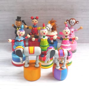 Può includere: Una collezione di giocattoli in legno colorati a tema circo, tra cui clown, un cavallo, una scimmia ed elefanti. I giocattoli sono dipinti con colori vivaci e poggiano su basi rotonde o cilindriche. I giocattoli hanno braccia e gambe mobili.