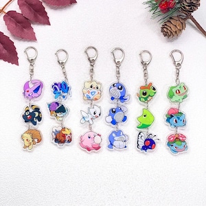 May include: Six vertical acrylic keychains, each featuring three distinct colourful cartoon character charms attached by silver metal chains and lobster clasps. The characters include various creatures in shades of pink, blue, green, yellow, and purple.