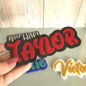 May include: A collection of name cutouts in various colors and styles. The name "Taylor" is prominently displayed in red, outlined in black, and the name "Michael" is in silver glitter. Other names include "Victoria" and "Annie".