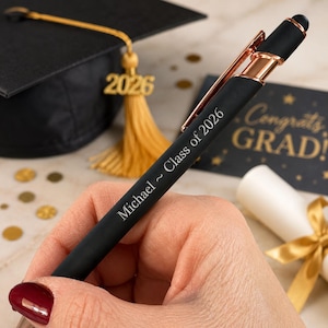 May include: A black pen with rose gold accents, engraved with "Michael ~ Class of 2026." The pen is held in a hand, with a graduation cap and a "Congrats Grad!" card in the background. Gold tassel and confetti complete the graduation theme.