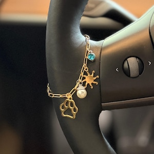 May include: Gold-toned charm bracelet with a paw print, sun, pearl, and blue crystal charms. The bracelet is attached to a black steering wheel. The charms are connected by a delicate chain.