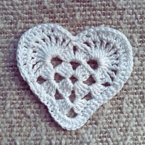 May include: A white, crocheted heart-shaped doily. The heart is made of white yarn and features an openwork design. The doily is placed on a textured, beige fabric background. The heart's dimensions are approximately 3 inches wide and 2.5 inches tall.