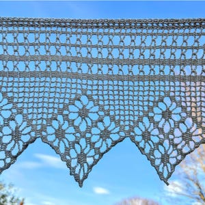 Puede incluir: Una cenefa de encaje de ganchillo gris con un diseño floral y geométrico. La cenefa tiene un borde festoneado y se muestra contra un cielo azul. El intrincado calado permite el paso de la luz, creando una apariencia delicada y aireada.