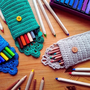 Op de afbeelding: Een verzameling kleurrijke kunstbenodigdheden, waaronder potloden, stiften en pastelkrijtjes, gerangschikt op een houten oppervlak. De afbeelding toont drie handgemaakte gehaakte etuis in blauw, groen en grijs, elk met een knoopsluiting en een decoratieve rand.