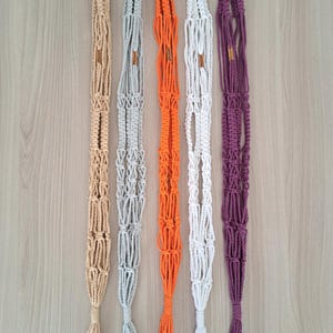 Puede incluir: Cinco maceteros de macramé en diferentes colores: beige, gris, naranja, blanco y morado. Cada macetero tiene un bucle en la parte superior y una borla en la parte inferior.
