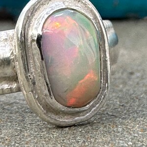 May include: A silver ring with an oval-shaped opal gemstone. The opal has a white base with flashes of pink, orange, and green.