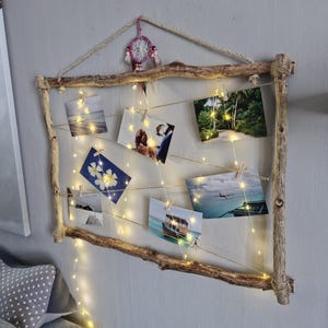 Peut inclure: Un présentoir photo rustique en bois avec des guirlandes lumineuses et des photos attachées. Le cadre est fait de branches naturelles, et les photos sont maintenues par de petites pinces en bois. Un attrape-rêves est suspendu en haut.