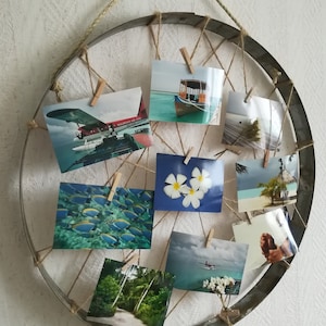 Könnte beinhalten: Ein rustikaler Holzkreis mit darübergespanntem Bindfaden, der verschiedene Fotos mit Wäscheklammern hält. Die Fotos zeigen tropische Szenen, darunter ein Boot, ein Flugzeug, Blumen und Fische.
