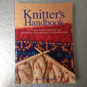 Può includere: Un libro intitolato "Knitter's Handbook" con copertina beige e testo blu. La copertina presenta primi piani di tessuto a maglia nei colori rosso, blu e crema, con ferri da maglia. Il libro è una guida completa per il lavoro a maglia a mano.