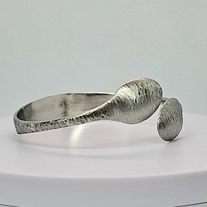 May include: A silver-toned open cuff bracelet featuring a textured, hammered finish. The design includes a wider, elongated oval end and a smaller, circular end, both with a matte, metallic surface that reflects light across the uneven metal texture.