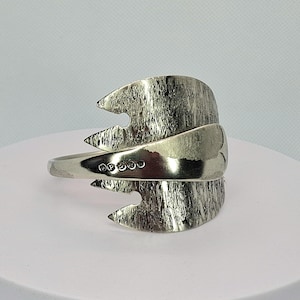 May include: A silver-toned metal cuff bracelet crafted from a repurposed fork. The design features a textured, jagged edge on the bowl of the fork and a smooth, curved handle that wraps around. Five small circular hallmarks are visible on the handle.