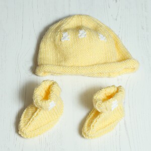May include: A knitted baby set in a soft yellow colour, including a beanie hat and two booties. The hat features three small white teddy bear appliqués. The booties also have white teddy bear appliqués.
