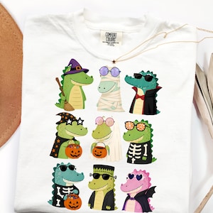 Puede incluir: Una camiseta blanca con una etiqueta Comfort Colors en el cuello, que presenta un estampado gráfico de nueve caimanes de dibujos animados vestidos con varios disfraces de Halloween, incluyendo una bruja, una momia, un vampiro y esqueletos.