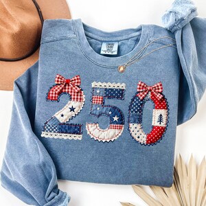 Op de afbeelding: Een lichtblauwe sweatshirt met het nummer "250" op de voorkant. De cijfers zijn gemaakt van verschillende rood-, wit- en blauwgepatroneerde stoffen, met een rood-witte ruitstrik bovenop de "2" en de "0".
