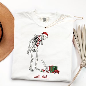 Könnte beinhalten: Ein weißes T-Shirt mit einer Grafik eines Skeletts, das eine rot-weiße Weihnachtsmütze und einen gestreiften Schal trägt. Das Skelett blickt auf seinen abgetrennten Arm, der nach einem grünen Geschenk mit roter Schleife und Ornamenten greift. Text: well, shit...