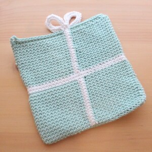 May include: A crocheted gift-shaped potholder in a light teal color, with white ribbon detailing. The potholder has a loop for hanging and is made of a textured yarn. The design resembles a wrapped present.