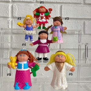 May include: Six Cabbage Patch Kids miniature figurines labeled #1 through #6. The figures include a red-haired child with a hammer, a blonde ballerina, a brown-haired angel, a dark-haired child in a purple dress, a red-haired child with a bear, and a blonde angel.