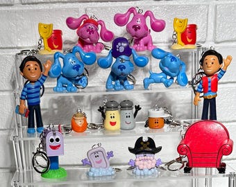 Könnte beinhalten: Eine Sammlung von Schlüsselanhängern und Figuren mit Charakteren, darunter Blue's Clues-Charaktere, ein roter Stuhl sowie Salz- und Pfefferstreuer, die auf einem transparenten, mehrstufigen Regal ausgestellt sind. Die Schlüsselanhänger weisen verschiedene Farben und Designs auf.