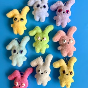 May include: Nine plush bunny keychains in various pastel colors, including yellow, blue, pink, and green, with expressive eyes and small metal chain attachments. The bunnies have a soft, fuzzy texture and are arranged on a bright blue background.