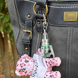 May include: A gray and black handbag with a decorative keychain. The keychain features a plush pink leopard with black spots, a white shirt, and a silver and green metal claw.