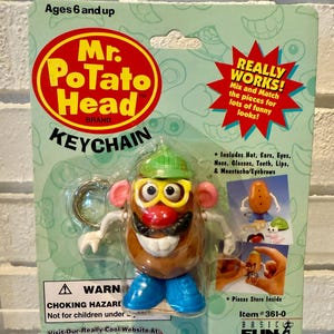May include: Mr. Potato Head keychain, a classic toy, packaged on a green and white card. The keychain features a brown potato-shaped body with interchangeable parts, including a hat, ears, eyes, nose, glasses, teeth, lips, and mustache. The packaging includes the text "Ages 6 and up."