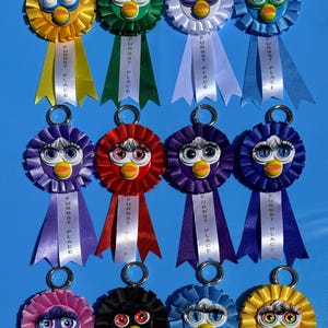 May include: A collection of colorful Furby-themed rosettes. Each rosette features a Furby face with large eyes and a beak, surrounded by ruffled ribbon. Ribbons with "FURBST PLACE" text hang below, attached to a metal ring.