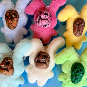May include: A collection of plush bunny figures in various colors, including white, pink, yellow, and green. Each bunny has a unique, detailed face attached. The figures are arranged on a blue surface.