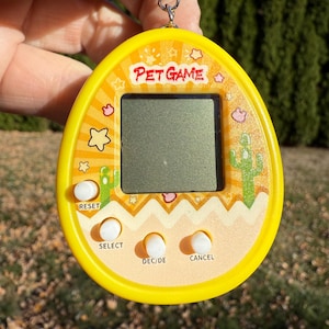 May include: Yellow Pet Game handheld electronic game with a keychain. The game features a small screen, buttons labeled "RESET", "SELECT", "DEC/DE", and "CANCEL". The design includes stars, clouds, and cactus graphics.
