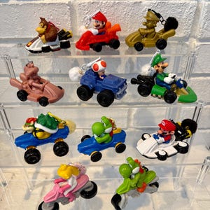 May include: A collection of miniature Mario Kart character figurines in various karts and motorcycles. The figurines feature characters like Mario, Luigi, and Yoshi, in a variety of colors including red, green, and blue. The figurines are displayed on clear shelves.