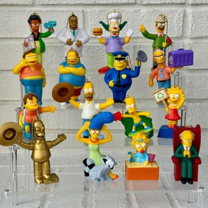 May include: A collection of colorful Simpsons character figurines, each with unique poses and outfits. The figurines include Homer, Marge, Bart, Lisa, and other characters from the animated TV show. They are displayed on a clear, tiered stand.