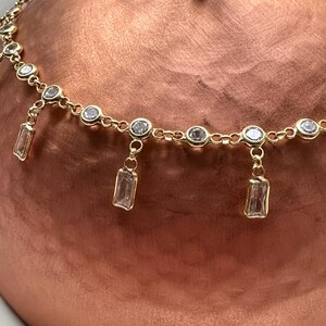 May include: A gold-toned necklace with a delicate chain design. The necklace features small, round, clear gemstones and rectangular, clear gemstone pendants. The jewelry is displayed against a copper-colored background.