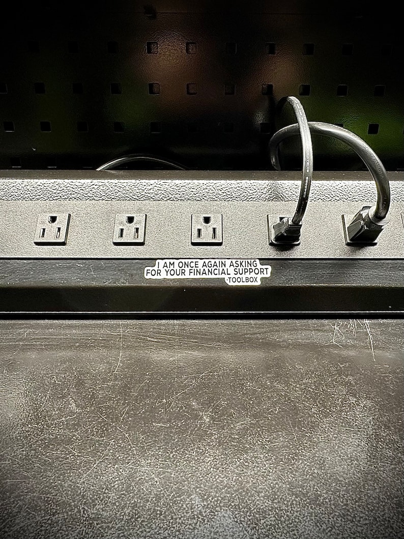 Puede incluir: Una regleta negra con m&uacute;ltiples enchufes y una pegatina que dice "I AM ONCE AGAIN ASKING FOR YOUR FINANCIAL SUPPORT TOOLBOX". Dos cables de alimentaci&oacute;n negros est&aacute;n enchufados.
