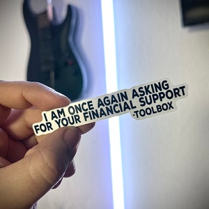 Puede incluir: Una pegatina blanca con texto negro que dice "I AM ONCE AGAIN ASKING FOR YOUR FINANCIAL SUPPORT -TOOLBOX". La pegatina se sostiene en una mano, con una guitarra negra borrosa en el fondo.