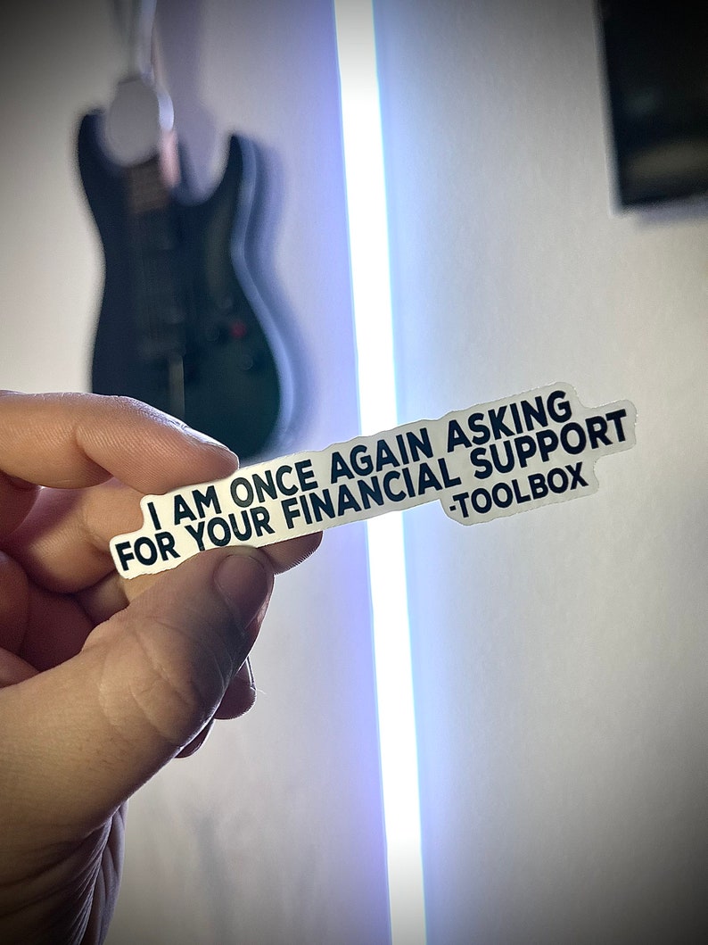 Puede incluir: Una pegatina blanca con texto negro que dice "I AM ONCE AGAIN ASKING FOR YOUR FINANCIAL SUPPORT -TOOLBOX". La pegatina se sostiene en una mano, con una guitarra negra borrosa en el fondo.