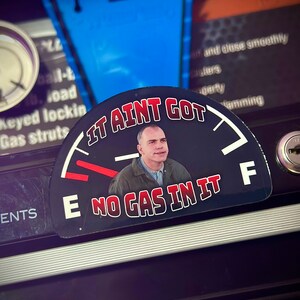 May include: A black and white gas gauge sticker with a red needle pointing to empty. The sticker features a man with a surprised expression and the text "It ain't got no gas in it".