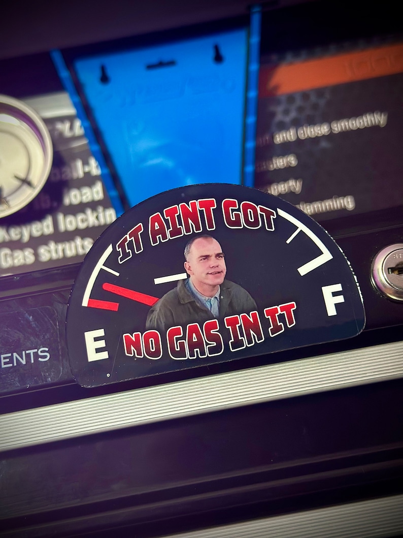 May include: A black and white gas gauge sticker with a red needle pointing to empty. The sticker features a man with a surprised expression and the text "It ain't got no gas in it".