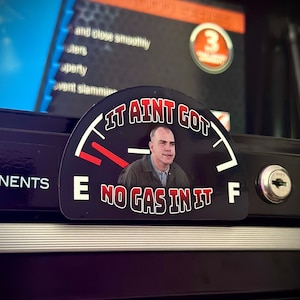 May include: A black and red gas gauge sticker with a white background. The gauge reads "It Ain't Got No Gas In It" and features a man's face in the center.