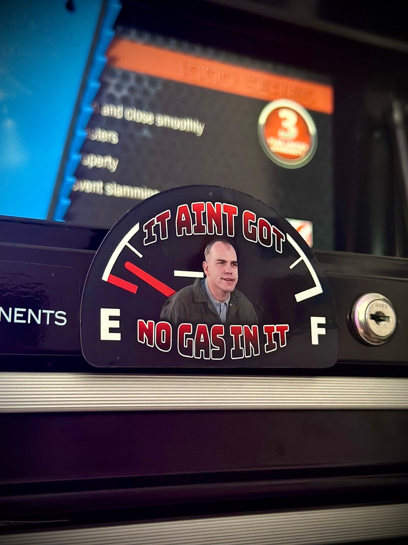 May include: A black and red gas gauge sticker with a white background. The gauge reads "It Ain't Got No Gas In It" and features a man's face in the center.