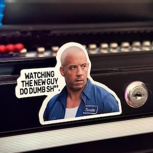 May include: A sticker featuring Vin Diesel as Dominic Toretto from the Fast and Furious franchise. The sticker has a speech bubble with the text "WATCHING THE NEW GUY DO DUMB SH**."
